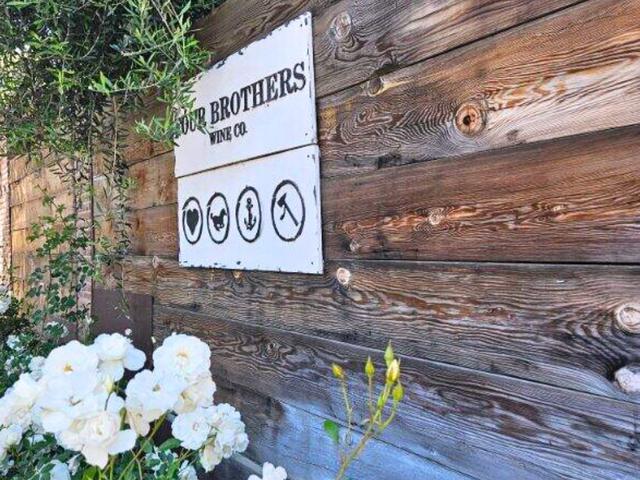 Weathered wooden wall with a white sign reading 'Four Brothers Wine Co.' adorned with icons of a heart, a rooster, an anchor, and a pickaxe, surrounded by white blooming flowers.