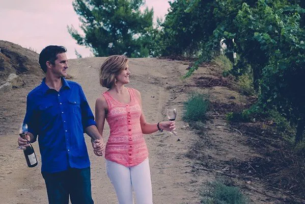 Couple walking through vineyard with wine bottle and glasses