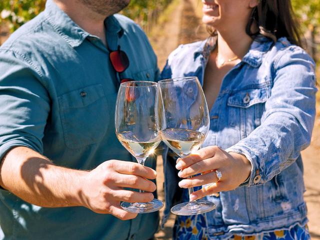 Couple toasting with white wine glasses in a vineyard, with the woman wearing a diamond engagement ring.