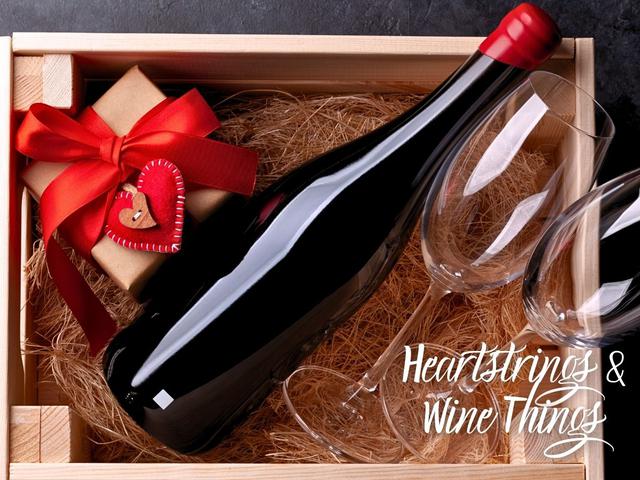 Wooden gift box containing a wine bottle, two wine glasses, and a small wrapped gift tied with a red ribbon and heart-shaped tag, with the title "Heartstrings & Wine Things."