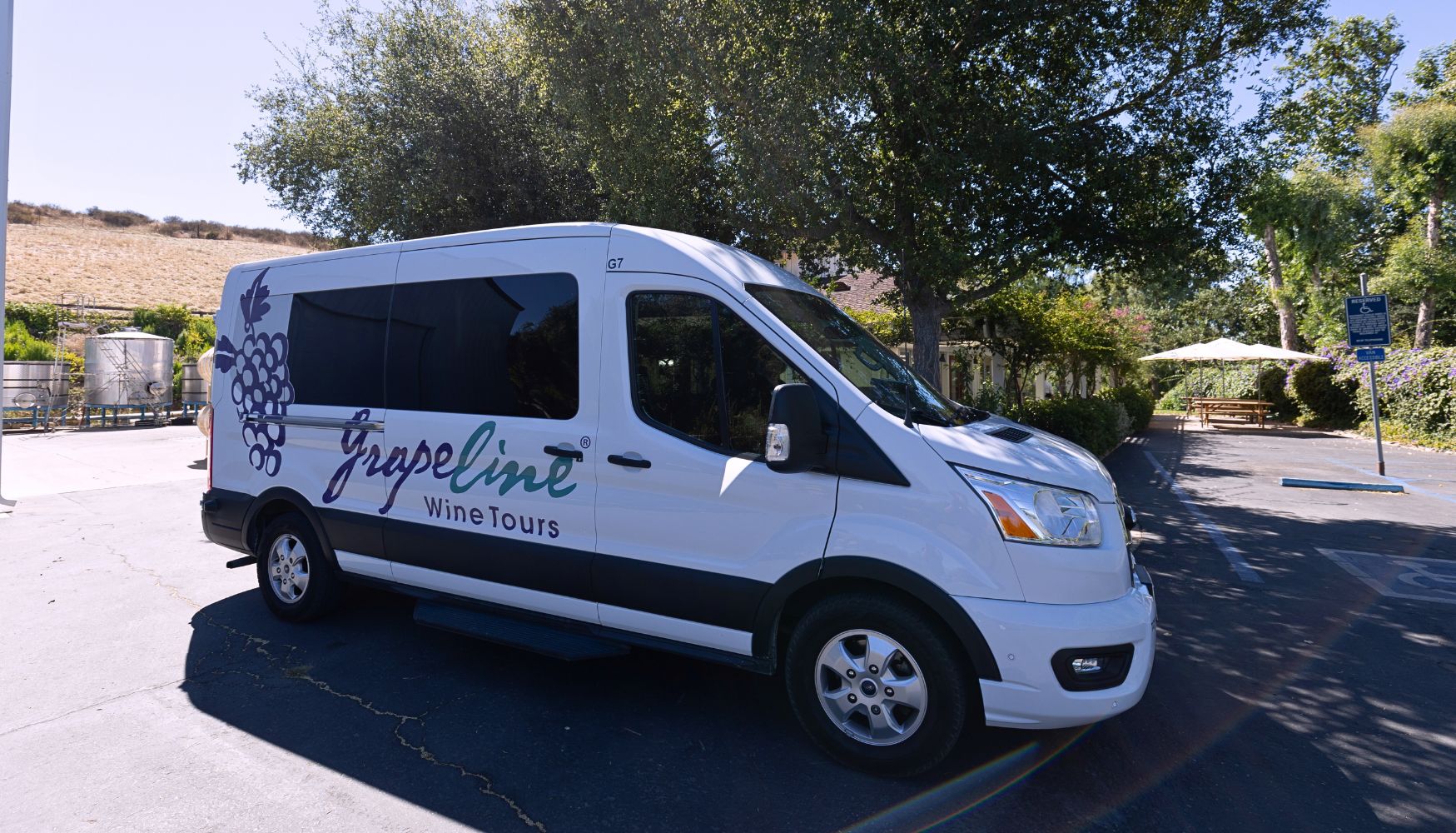 White Grapeline Wine Tours van parked outside a vineyard, surrounded by trees and rolling hills on a sunny day.