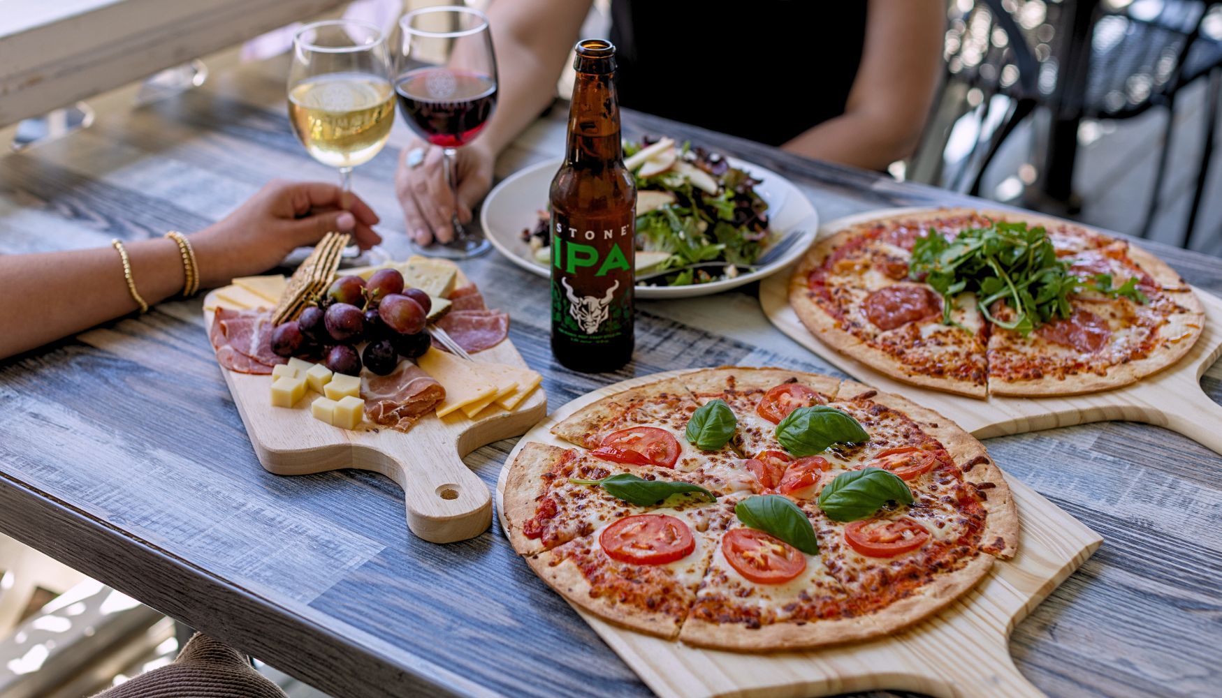 Couple girlfriends enjoying wood-fired pizzas, charcuterie board, and fresh salad paired with glasses of wine and a craft beer on an outdoor table.