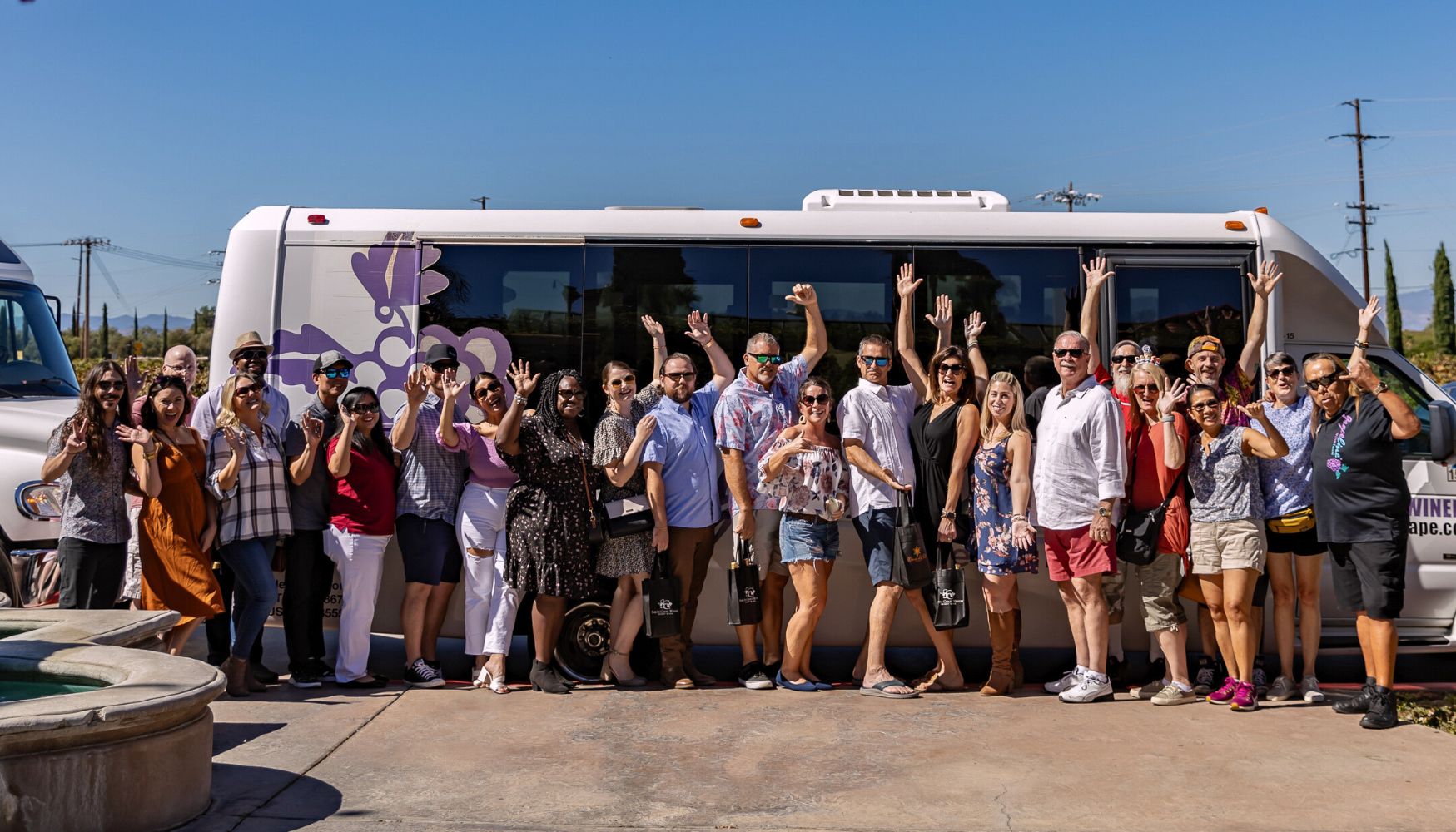 Large group posing in front of a wine tour bus, smiling and raising their hands in celebration.