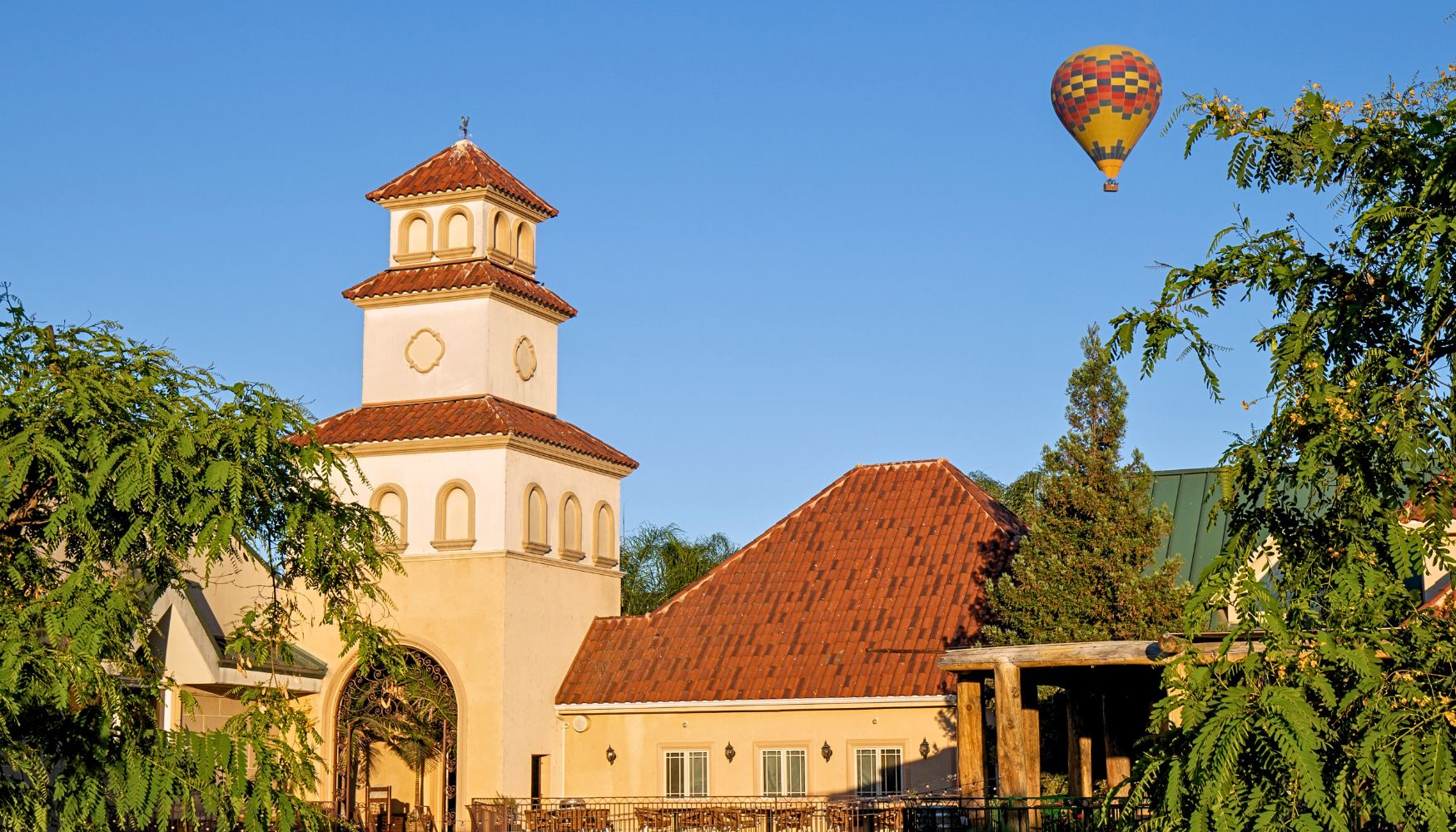 Hot air balloon floating over Temecula vineyards with a Mediterranean-style winery building in the foreground.