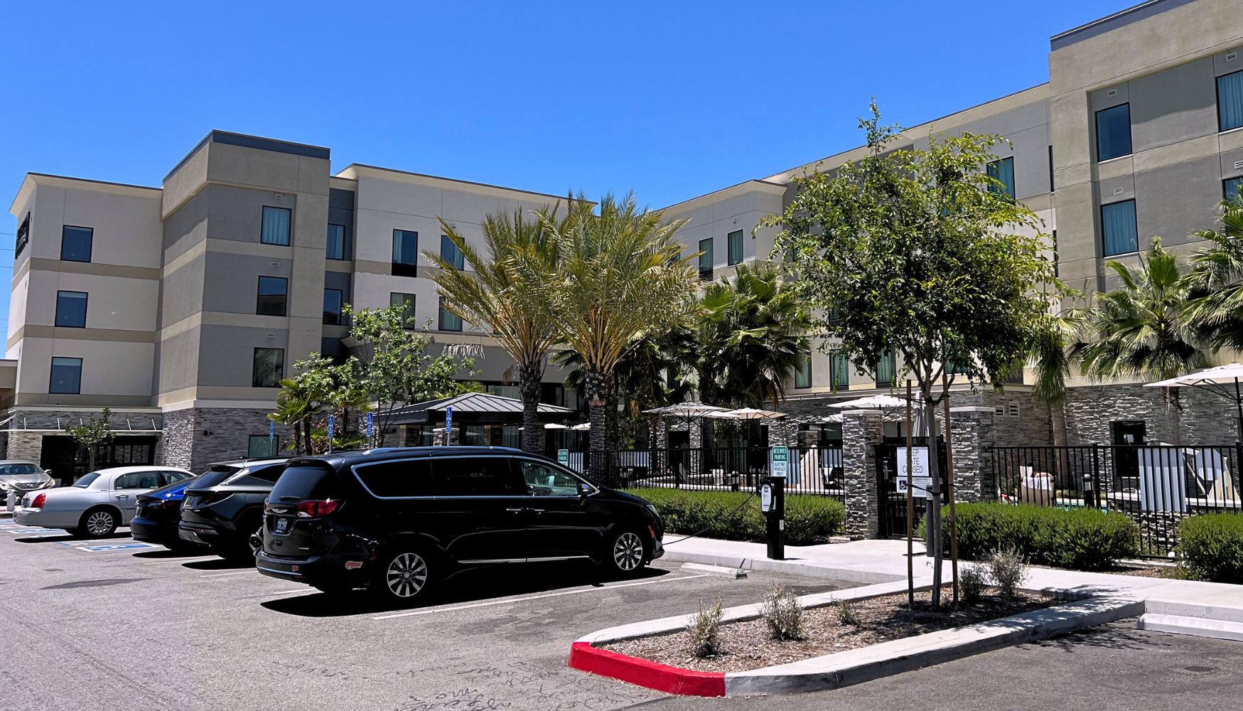 Modern hotel in Temecula with palm trees, a fenced pool area, and parked cars under a clear blue sky.