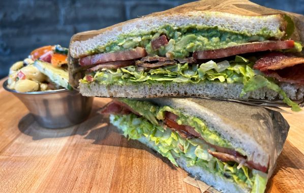 A hearty BLTA sandwich featuring bacon, fresh lettuce, ripe tomato slices, and avocado spread, all layered between two slices of sourdough bread. The sandwich is served with a side of rustic macaroni salad, providing a satisfying and flavorful meal.