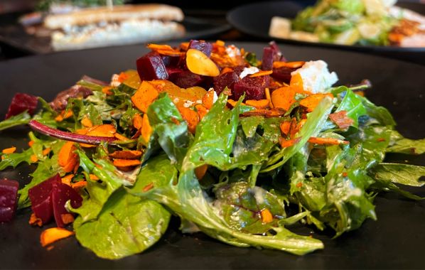 A vibrant, roasted beet salad featuring a mix of fresh greens, diced beets, toasted spiced almonds, and smoked goat cheese, all lightly dressed in a Green Goddess Dressing and served on a black plate.