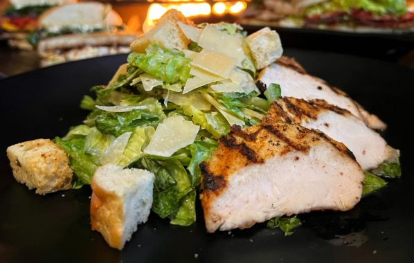 A classic Chicken Caesar salad featuring tender, grilled chicken arranged over a bed of crisp romaine lettuce, topped with crunchy croutons, shaved Parmesan cheese, and a creamy Caesar dressing, creating a perfect balance of flavors and textures.