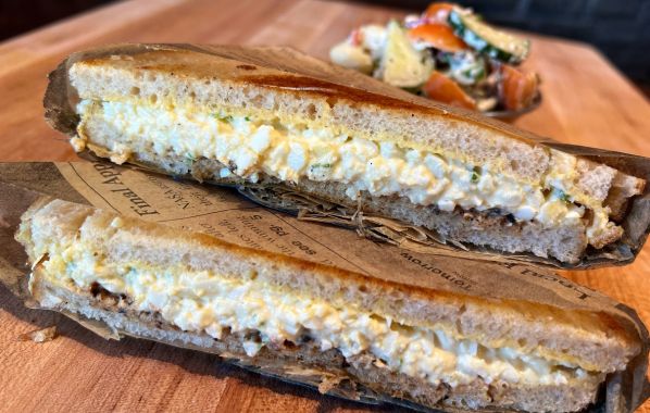 This egg salad sandwich features a generous filling of creamy, well-seasoned egg salad, tomato jam, and parsley, sandwiched between two slices of sourdough bread. It's served with a side of rustic macaroni salad, offering a satisfying and comforting meal with classic flavors.