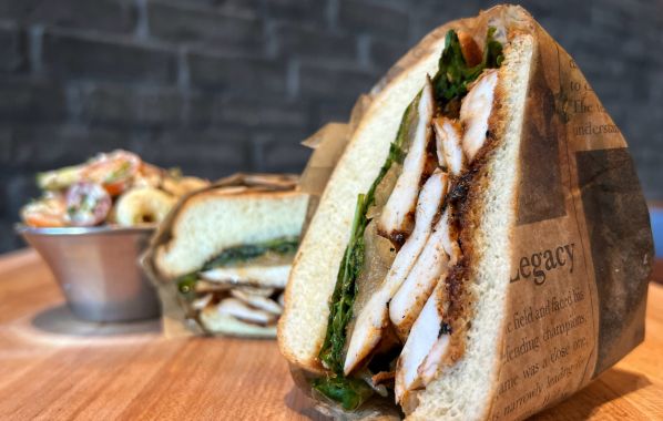 The grilled chicken sandwich features tender, juicy slices of chicken, expertly grilled, layered with fresh baby arugula, carmalized nion, tomato jam, and balsamic glaze on a brioche bun. It comes with a side of rustic macaroni salad, creating a wholesome lunch.